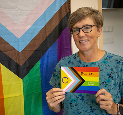 Gail smiling in front of Pride flag holding a Pride postcard