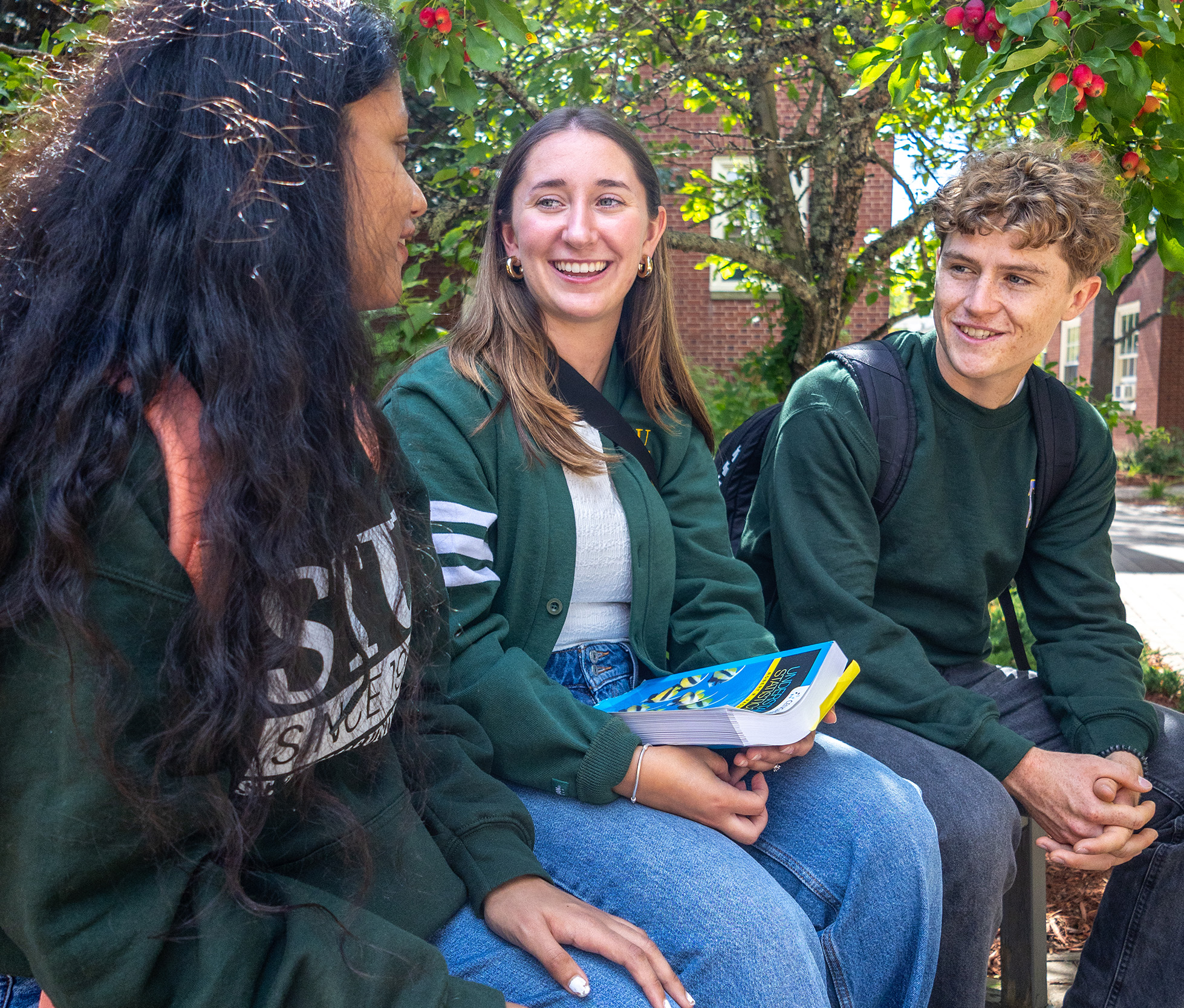 Three students sit on a bench outside on campus