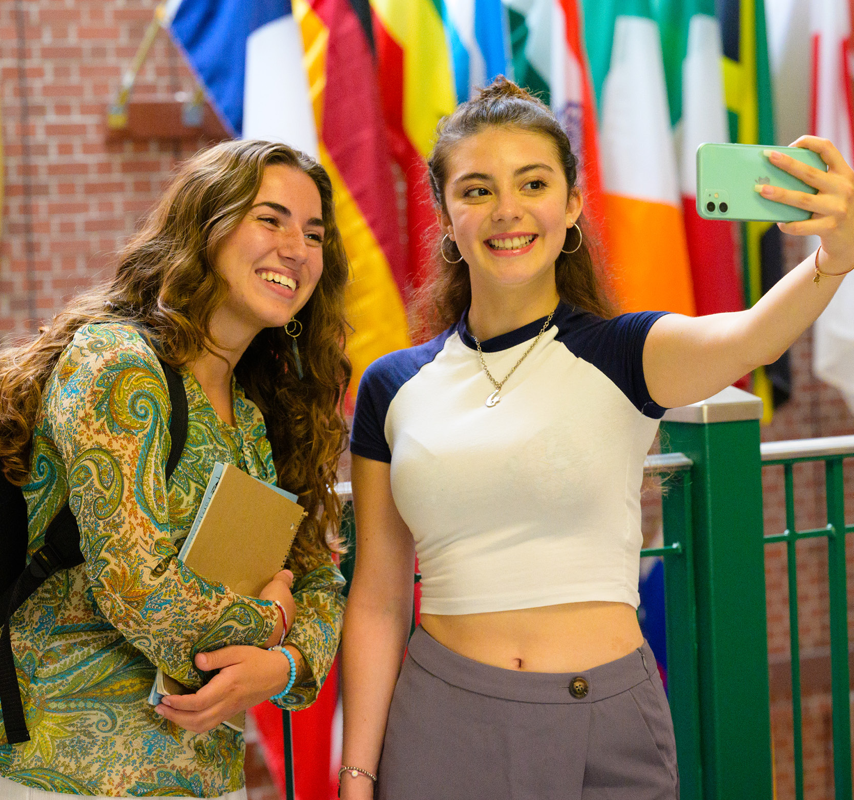 Two students posing for a selfie in front of a wall of flags