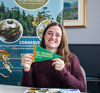 Student at internship office sitting at desk holding STU pennant, next to Nature Trust NB banner