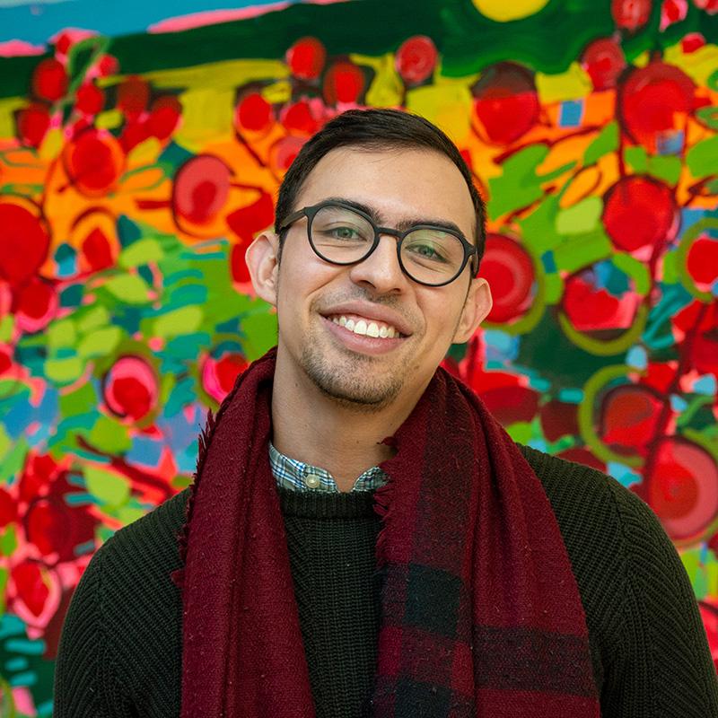 A man wearing glasses and red scarf stands in front of a colorful painting