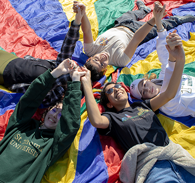Group of student volunteers laying down on rainbow mat outdoors smiling holding hands in the air