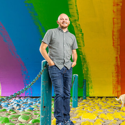 A man wearing a grey short sleeve button down shirt and jeans standing in front of a rainbow mural