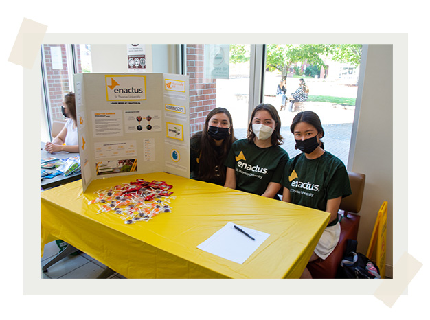 Three students from the Enactus STU club sit at a yellow booth in James Dunn Hall